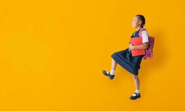 Asiatisches Studentenkind Mädchen trägt Schuluniform und hält Buch mit Gehen isoliert auf gelbem Hintergrund mit Beschneidungspfaden für Designarbeiten leerer freier Raum