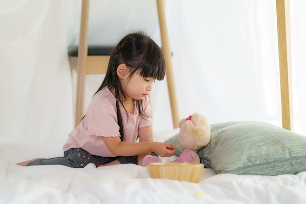 Asiatisches süßes kleines Mädchen spielen ihre Puppe und Fütterungssnack, während sie in einer Deckenfestung im Wohnzimmer zu Hause sitzen