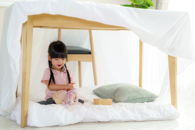 Asiatisches süßes kleines mädchen spielen ihre puppe, während sie in einer deckenfestung im wohnzimmer zu hause sitzen