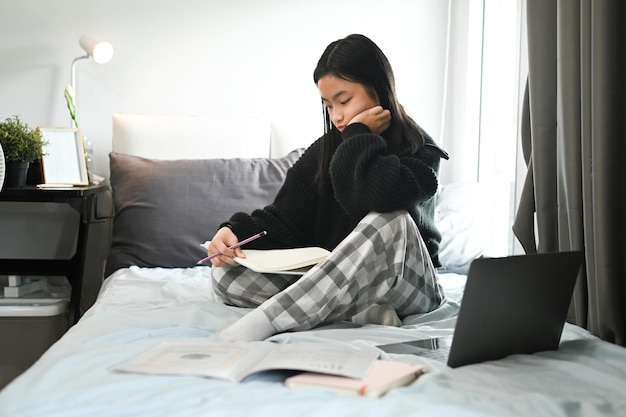 Asiatisches Teenagermädchen sitzt auf dem Bett und macht Hausaufgaben. Fernunterricht studiert Online-Konzept