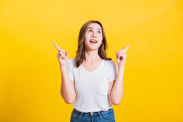 Asiatisches thailändisches glückliches Porträt, schöne, süße junge Frau, die ein weißes T-Shirt trägt, macht Geste, zwei Finger zeigen nach oben, oben schauend, Studioaufnahme isoliert auf gelbem Hintergrund mit Kopierraum