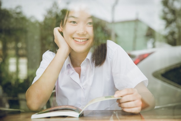 Asiatisches Universitätsmädchen jugendlich süß lächelnd durch das Glasfenster schauen Schatten reflektieren den Effekt beim Sitzen beim Lesen eines Buches