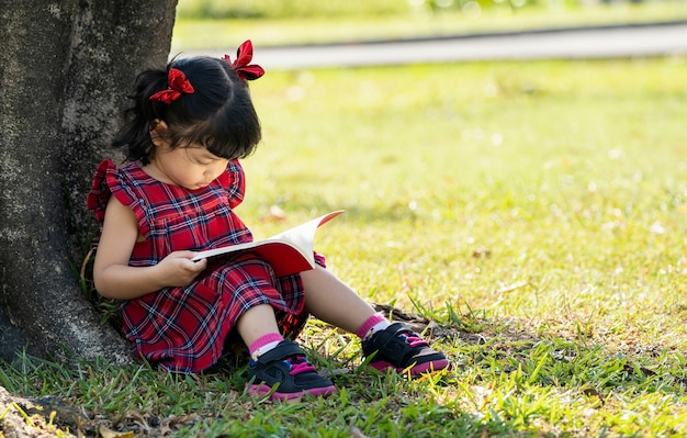 Asiatisches Vorschulmädchen, das das Buch unter dem Baum liest