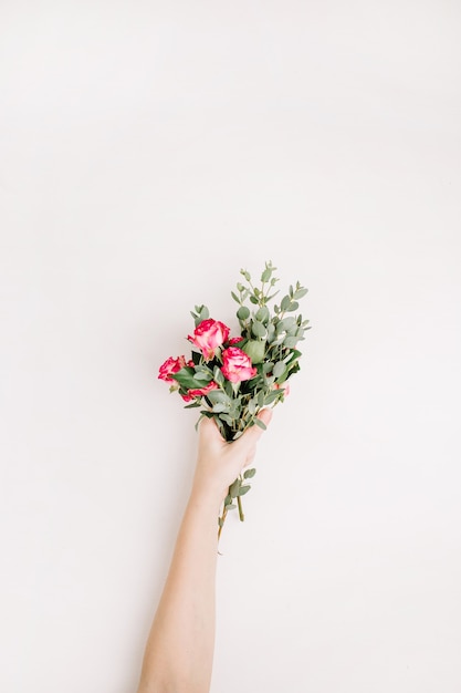 Asimiento de la mano de la mujer ramo de flores color de rosa y eucalipto sobre fondo blanco. Endecha plana, vista superior