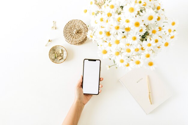 Asimiento de la mano de las mujeres smartphone con pantalla en blanco. Espacio de trabajo de escritorio de oficina en casa con ramo de flores de manzanilla margarita y cuaderno en blanco