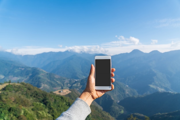 Asimiento de la mano del teléfono móvil en las montañas y el paisaje de campo y el cielo azul con nubes