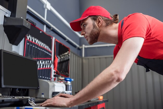 Asistencia de información. Trabajador de taller adulto joven enfocado atento en gorra roja y camiseta mirando interesado en la computadora