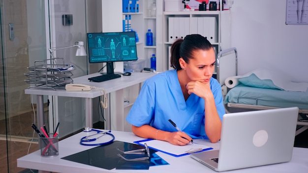 Asistente agotado mirando la pantalla del portátil, escribiendo en el portapapeles, trabajando desde la oficina del hospital. Médico en medicina uniforme escribiendo una lista de pacientes consultados y diagnosticados, realizando una investigación.