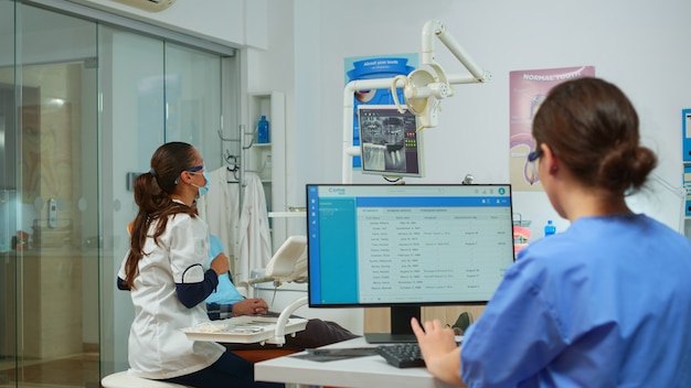 Asistente de dentista haciendo citas en la computadora mientras el médico de odontología apunta en la pantalla digital que muestra los implantes dentales. Estomatólogo explicando la radiografía de los dientes en el monitor de la clínica estomatológica.