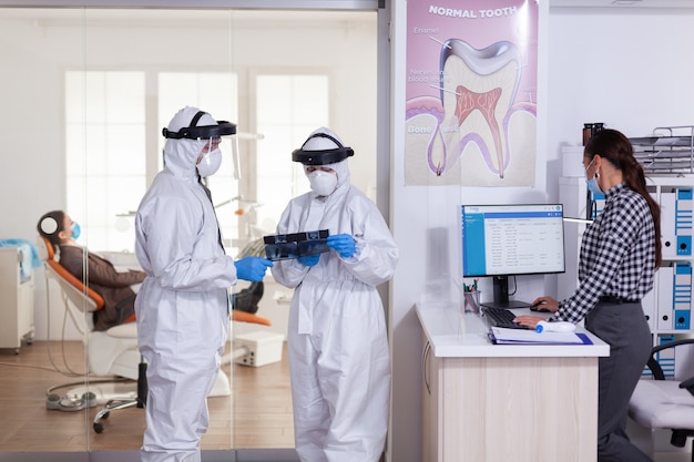 Asistente de estomatología vistiendo traje de ppe shiled facial como precación de seguridad con el dentista discutiendo radiografías en la sala de espera de la clínica dental