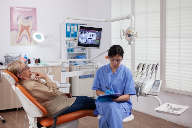 Asistente en el gabinete del dentista durante el control de higiene bucal de la anciana sentado en una silla. Mujer mayor hablando con una enfermera médica en la oficina de estomatología sobre el problema de los dientes.