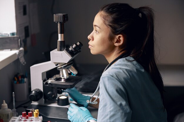 Asistente de laboratorio asiático pensativo se sienta cerca del microscopio mirando por la ventana en el lugar de trabajo