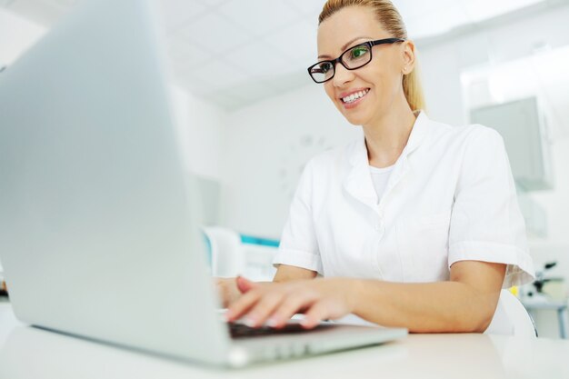 Asistente de laboratorio en bata con anteojos sentado en el laboratorio y escribiendo en el informe de la computadora portátil.