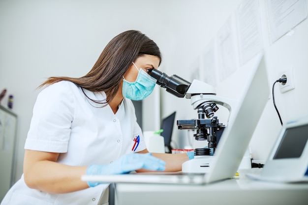 Foto asistente de laboratorio caucásico encantador en uniforme blanco, con máscara protectora y guantes de goma sentado en el laboratorio mirando microscopio a través de muestra de sangre y usando una computadora portátil.