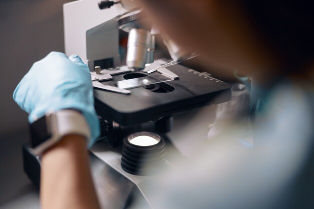 Asistente de laboratorio en guantes pone la muestra de material en un portaobjetos en el microscopio que trabaja en el laboratorio