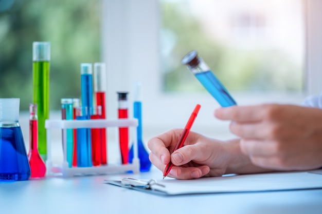 El asistente de laboratorio realiza investigaciones clínicas de laboratorio, sostiene un tubo de ensayo líquido azul en sus manos y escribe el resultado del estudio en el portapapeles. Concepto de medicina, farmacia y cosmetología.