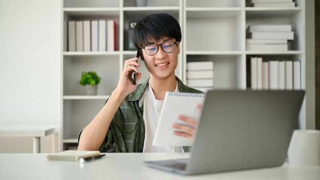 Un asistente masculino asiático inteligente miró los detalles en un bloc de notas mientras hablaba por teléfono