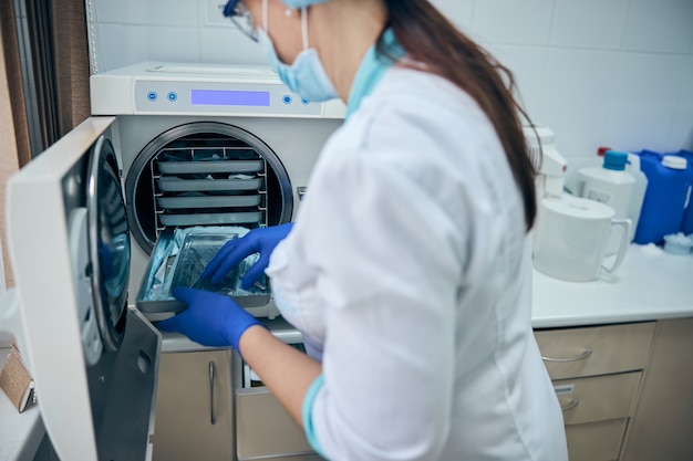 Asistente médico en guantes azules esterilizar herramientas de acero dental en autoclave