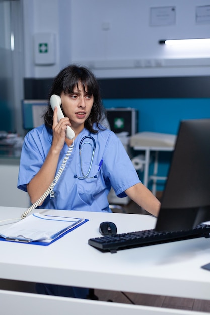 Asistente médico hablando por teléfono para cita de chequeo, trabajando hasta tarde. Enfermera mediante teléfono fijo para comunicación remota en línea con el paciente en el gabinete de atención médica.