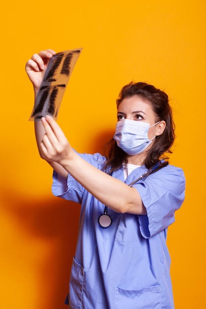 Asistente médico con mascarilla y mirando rayos X para encontrar el diagnóstico. Enfermera con protección contra virus y uniforme con estetoscopio mientras mira radiografía