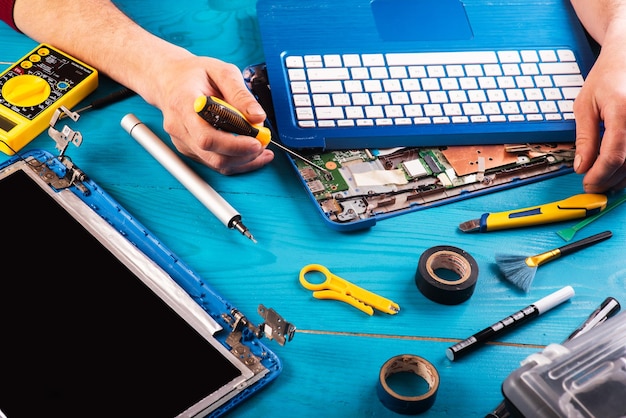 El asistente repara la computadora portátil con herramientas y manos en la vista superior de la mesa de madera azul