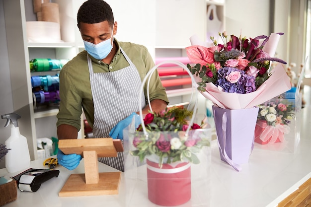 Asistente de tienda de flores en guantes y mascarilla operando una tableta acostada sobre un soporte de madera