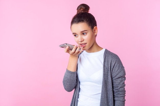 Asistente virtual. Retrato de una adolescente morena con peinado de moño con ropa informal hablando por teléfono usando una aplicación de voz digital para buscar datos en Internet. tiro del estudio aislado sobre fondo de color rosa