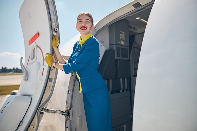 Asistente de vuelo hermosa joven mirando a la cámara y sonriendo mientras coloca las manos en la puerta del avión