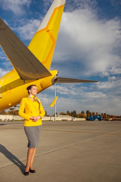 Asistente de vuelo de mujer alegre en uniforme de trabajo de aviación de pie cerca del avión en el aeropuerto