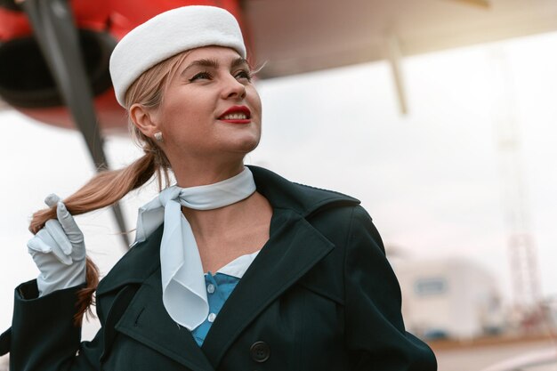 Asistente de vuelo de mujer en uniforme de pie cerca del avión en el aeropuerto y mirando hacia otro lado