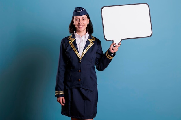 Asistente de vuelo sonriente sosteniendo una burbuja de habla en blanco, azafata de pie con un marco de comunicación simulado. Azafata en uniforme con globo de mensaje vacío, lugar para texto