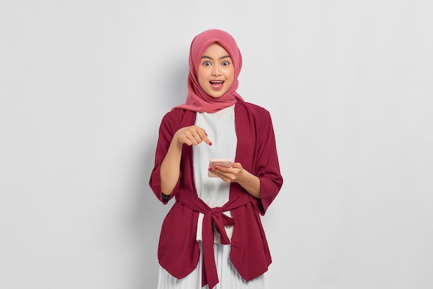 Asombrada hermosa mujer asiática con camisa informal y hiyab señalando con el dedo el teléfono móvil aislado sobre fondo blanco. Concepto de estilo de vida religioso de personas
