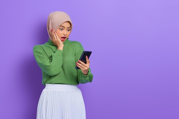 Asombrada hermosa mujer asiática con suéter verde e hiyab usando un teléfono móvil, leyendo un mensaje de buenas noticias aislado sobre un fondo morado