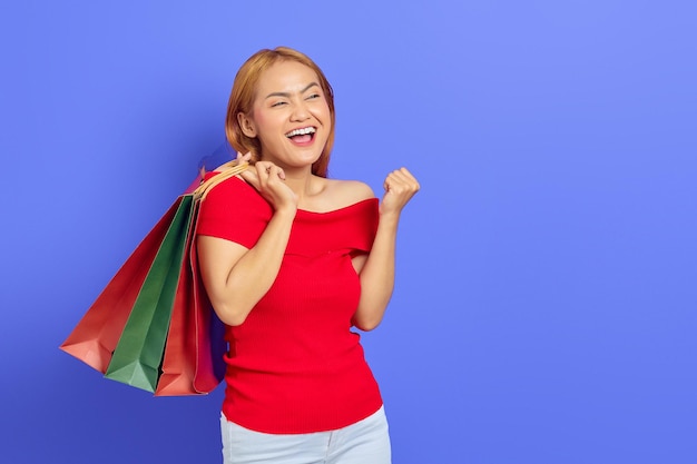 Asombrada hermosa mujer asiática vestida de rojo sosteniendo bolsas de compras, mirando el espacio de copia y celebrando la suerte, mostrando ofertas en línea aisladas sobre fondo morado