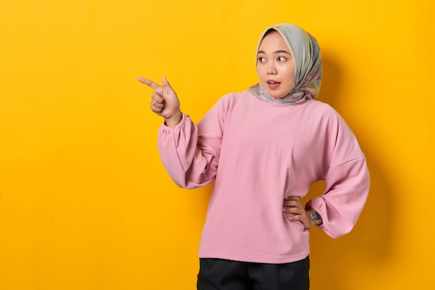 Asombrada joven asiática con camisa rosa señalando con el dedo el espacio de la copia con fondo amarillo