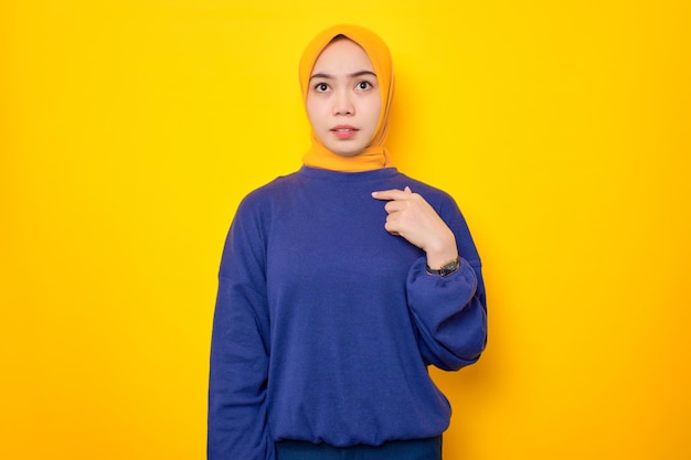 Asombrada joven musulmana asiática vestida con un suéter informal apuntándose a sí misma con la cara perpleja aislada sobre el fondo amarillo