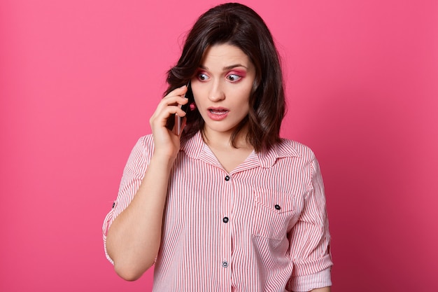 Asombrada mujer con atuendo casual hablando con alguien a través de un teléfono inteligente moderno, habiendo sorprendido la expresión facial, de pie con los ojos grandes contra la pared de rosas.