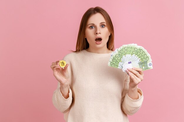 Asombrada mujer sorprendida sosteniendo una moneda de oro de la moneda criptográfica y un gran fanático de los billetes en euros mirando a la cámara con la boca abierta usando suéter blanco Foto de estudio interior aislada en fondo rosa