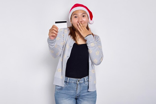 Asombrada y satisfecha joven asiática con sombrero de Navidad mostrando una tarjeta de crédito blanca en blanco en la cámara cubriendo la boca con la mano aislada en el fondo blanco