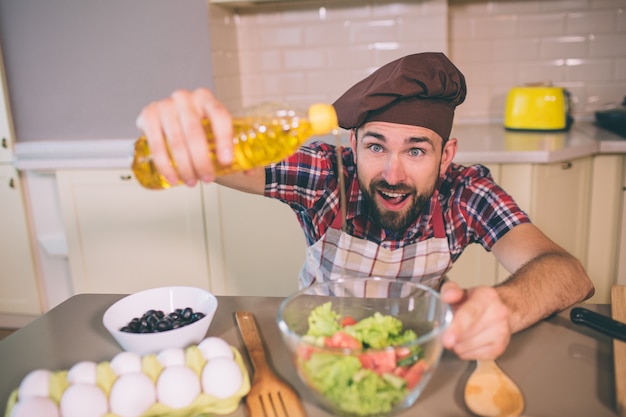 Foto asombrado y feliz chico se para a la mesa en la cocina y sostiene una botella de aceite. vierte un poco en un tazón con ensalada. guy mantiene la boca abierta.