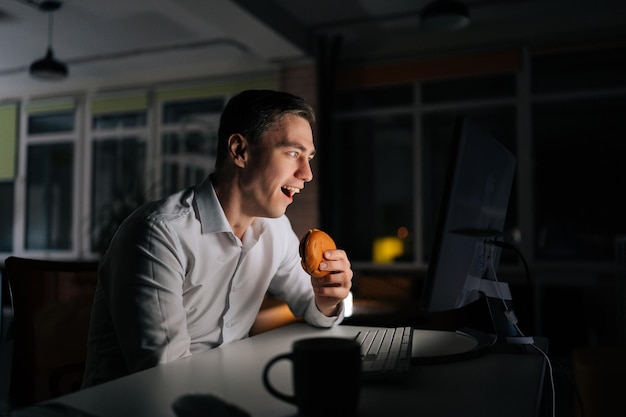 Asombrado freelance hombre hamburguesa para llevar mientras trabaja horas extras feliz emocionado mirando al escritorio