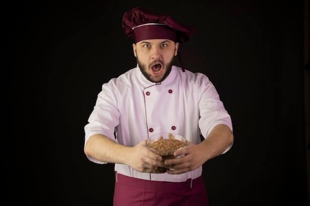 Asombrado joven chef barbudo en uniforme sosteniendo tazón de cereal