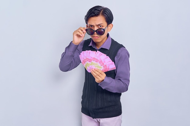 Asombrado joven empresario asiático sosteniendo dinero en efectivo en billetes de rupias y quitándose gafas aisladas sobre fondo blanco Concepto de estilo de vida de la gente