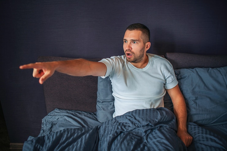 Foto asombrado y maravillado joven sentado en la cama en el dormitorio y mira a la izquierda. señala con el dedo. guy está excitado.