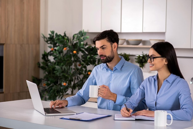 Aspecto inteligente Cintura arriba de alegres jóvenes colegas de negocios sentados en la cocina y usando una computadora portátil juntos