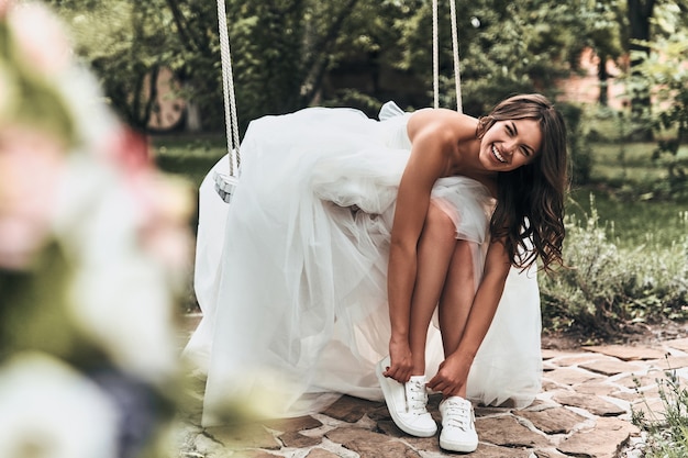 ¡Aspecto perfecto! Atractiva mujer joven en vestido de novia poniéndose zapatos deportivos y sonriendo mientras está sentado en el columpio al aire libre