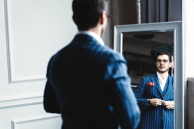 Aspecto perfecto. Reflexión de un joven apuesto en traje completo ajustando su chaqueta mientras está de pie frente al espejo en el interior.