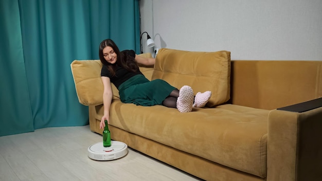 La aspiradora robot blanca entrega una botella de cerveza de alcohol a la  mujer, sentada en el sofá en la sala de estar. | Foto Premium