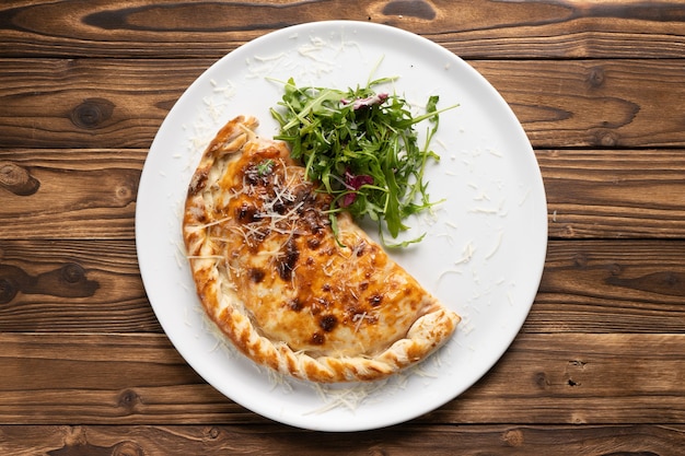 Assado para um calzone de pizza marrom crocante com rúcula fresca e queijo parmesão em um prato de cerâmica branca sobre uma mesa de cozinha de madeira.