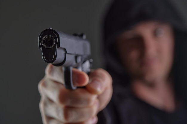 Assaltante com arma apontada para a câmera Homem de capuz ameaça com arma de fogo Arma nas mãos da pessoa Assassino ou ladrão armado Criminoso com pistola